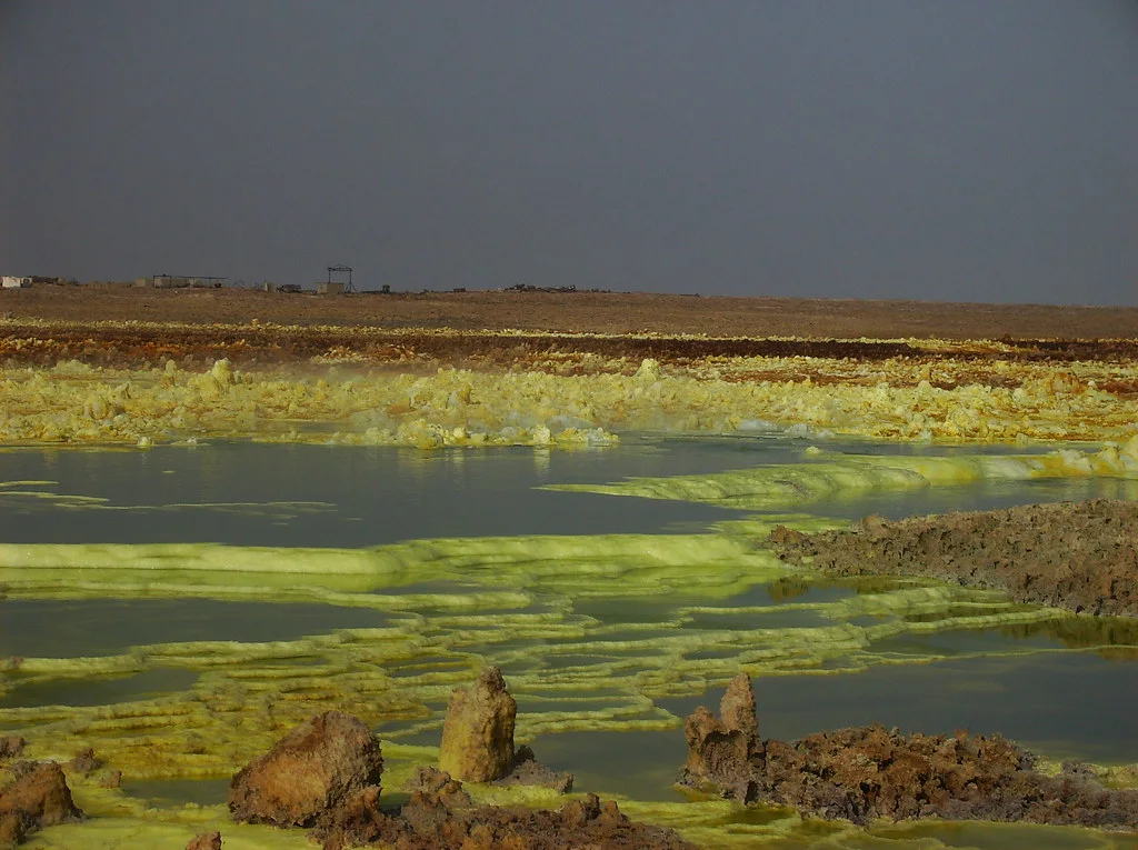Acid-green and sulphur-yellow mineral pools spread across the Dallol hydrothermal field beneath a dark grey sky, with brown salt pillars in the foreground.
