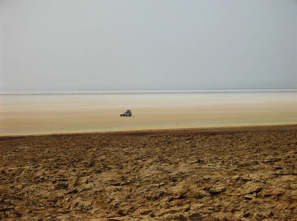 A lone 4x4 crosses the pale salt plain of the Danakil Depression beyond a band of brown crust.